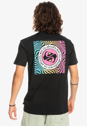 Mann mit schulterlangem lockigem Haar, der ein schwarzes T-Shirt mit farbigem wellenförmigem quadratischem und rundem Quiksilver-Logo auf dem Rücken trägt.