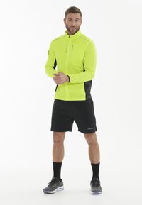 Herretøj til aktiv livsstil med en lys gul jakke med sorte detaljer, lynlåslommer, parret med sorte shorts og mørke sportssko.