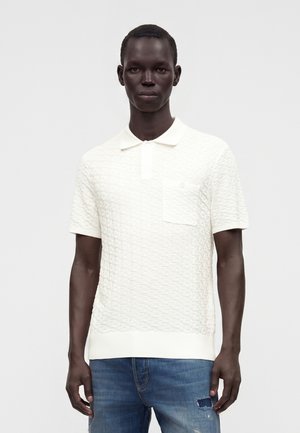 Homme portant un polo blanc texturé à manches courtes avec une poche poitrine et un jean bleu déchiré, debout devant un fond uni.