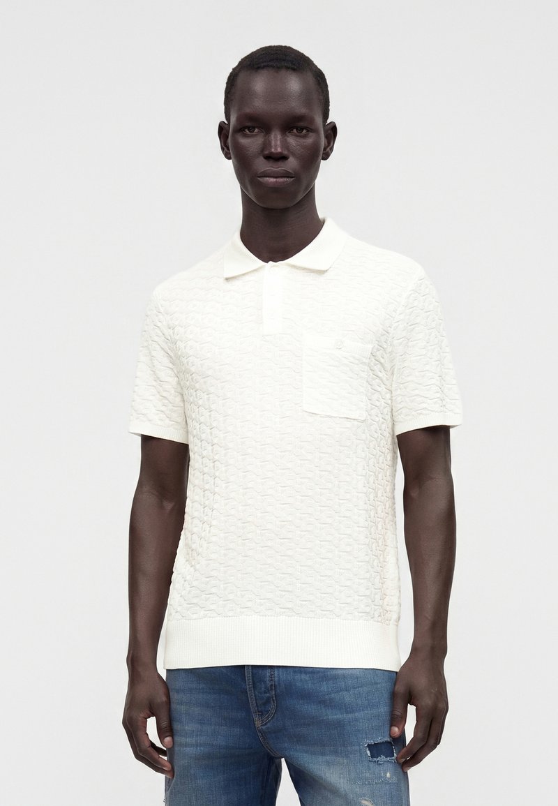 Homme portant un polo blanc texturé à manches courtes avec une poche poitrine et un jean bleu déchiré, debout devant un fond uni.