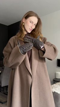 Γυναίκα με μπεζ παλτό και μαύρα γάντια ποζάρει μέσα σε εσωτερικό χώρο, με ανοιχτό καστανό μαλλί, φορώντας χρυσά σκουλαρίκια και ρολόι.