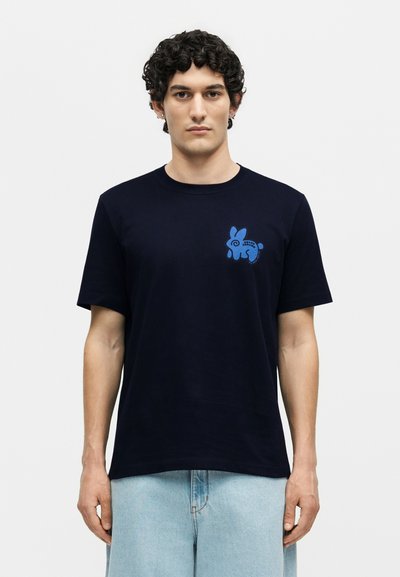 Νέος άνδρας με σγουρά μαλλιά που φοράει μαύρη μπλούζα με ένα μικρό μπλε σχέδιο ενός στυλιζαρισμένου κουνελιού στο αριστερό μέρος του στήθους.