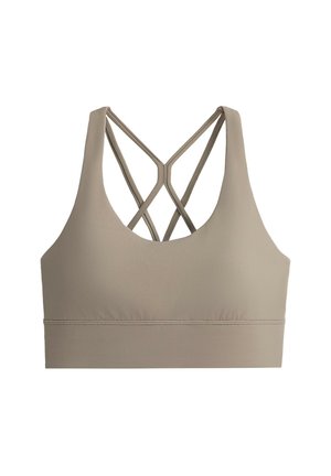 Femme portant un soutien-gorge de sport beige, un pantalon de jogging blanc, des baskets et une casquette blanche, debout les mains dans les poches devant un fond gris uni.