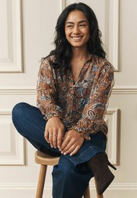 Hnedá blúzka s kvetinovým vzorom a volánovými rukávmi, spárovaná s tmavomodrými džínsami a hnedými čižmami na podpätku, model sediaci na drevenom stoličke.