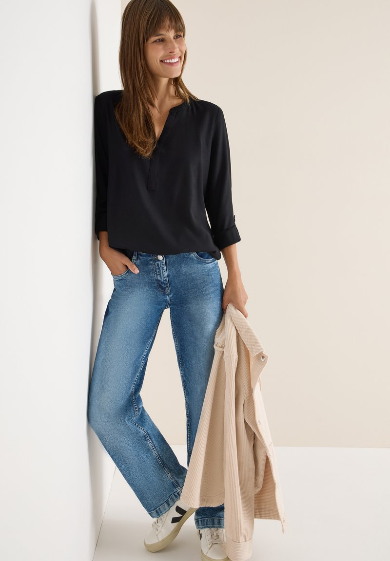 Sort V-hals skjorte med opsmøget ærmer, lyseblå jeans og hvide sneakers. Beige corduroyjakke holdt i hånden. Enkel, afslappet påklædning.
