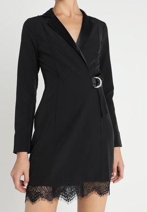 Czarna dopasowana sukienka żakietowa z długim rękawem, z satynowymi klapami, paskiem w talii i koronkowym wykończeniem na dole, noszona przez stojącą osobę na białym tle.