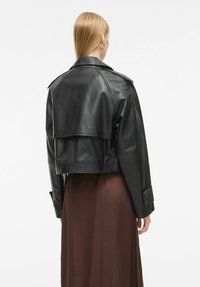 Juodas odinis trumpas švarkas su atvartų detale nugaros dalyje, mygtukų dekoracijomis ant rankovių ir lygiu paviršiumi. Dėvimas virš rudos suknelės.