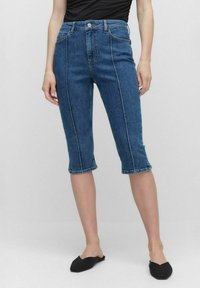 Un pantalon capri en denim bleu au waist haut, avec un design à cinq poches et des coutures contrastées. L'ourlet est légèrement retroussé.