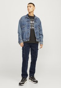 Chaqueta de mezclilla con un acabado azul desgastado, camiseta gráfica negra, pantalones cargo azul marino con bolsillos, y zapatillas deportivas negras con detalles grises.