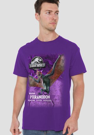 Mann trägt ein lila Jurassic World T-Shirt mit großem Pteranodon-Grafik und Arteninfo auf der Vorderseite vor einfarbigem Hintergrund.