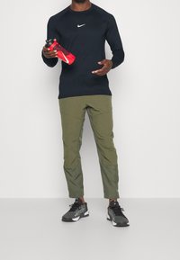Černé tričko s dlouhým rukávem Nike, olivové kalhoty se stranovými panely, držící červenou lahev na vodu a obuté černé a šedé sportovní boty.