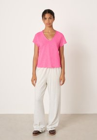 Ženska stoji ob beli steni, oblečena v rožnato majico z V-izrezom in kratkimi rokavi, svetlo sive široke hlače in temno rjave čevlje.