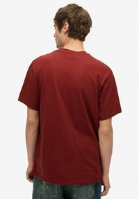 Ung mand med kort brunt hår iført en ensfarvet rød kortærmet T-shirt og blå jeans, set fra baggrunden mod en hvid baggrund.