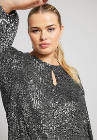 Top nero con paillettes, scollatura rotonda e dettaglio a chiave. Maniche lunghe, texture scintillante e vestibilità ampia. Orecchini pendenti argentati.