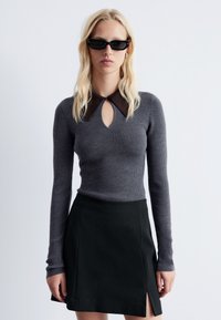 Grauer gerippter Pullover mit einem braunen Kragen und einem Schlitzdetail, kombiniert mit einem schwarzen Rock mit einem Seitenschlitz. Elegantes, tailliertes Design.