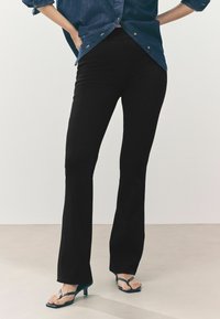 Zwarte flared broeken van rekbaar materiaal, met een hoge tailleband en een gladde textuur, in combinatie met blauwe hakslippers.