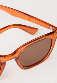 Orange transparente Sonnenbrille mit runden Rahmen. Braune getönte Gläser und glänzende Oberfläche. "SEASALT" auf dem Bügel gedruckt.
