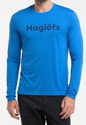 Mann trägt ein hellblaues Langarmshirt mit schwarzem "Haglöfs"-Logo, kombiniert mit schwarzer Hose, steht vor weißem Hintergrund.