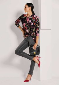 Schwarzes Oberteil mit Blumenmuster, graue Jeans, rote spitze High Heels; schwarze Umhängetasche, goldene Details, glatte Texturen. Modell posiert vor einer Wand.