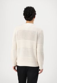 Cárdigan de punto color crema con rayas horizontales texturizadas, puños y dobladillo acanalados. El patrón de tejido abierto permite una buena transpirabilidad y opciones para superponer.