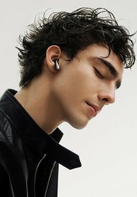 Kabellose Ohrstöpsel in einem eleganten schwarzen Design, teilweise sichtbar im Ohr einer Person mit lockigem Haar, die eine schwarze Lederjacke trägt.