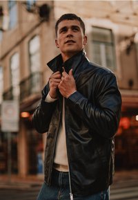 Black leather jacket with a high collar, featuring a zip closure and textured surface, worn over a light sweater and blue jeans.