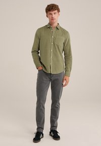 Camicia verde oliva con bottoni e tasca, abbinata a jeans grigi skinny e sneakers nere con strisce bianche. Stile casual, tessuto testurizzato.