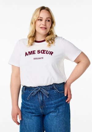 Bela majica s kratkimi rokavi in maroon krožnim izrezom, na kateri je v boldu natisnjeno "ÂME SØEUR SOULMATÉ". Ujemajoči se visoko pasjeni modri jeans.