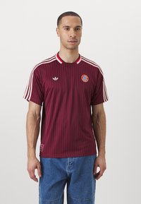 Maglia da calcio a righe bordeaux con dettagli bianchi, colletto a costine, logo Adidas e emblema del Bayern Monaco sul petto. Maniche corte.