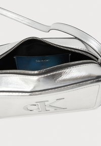 Strieborná kovová crossbody taška Calvin Klein so zipsom, hladkou textúrou a modrým interiérom s embosovaným logom.