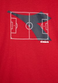 Červené tričko s bílou grafikou fotbalového hřiště, šedým trojúhelníkovým akcentem a logem "diadora" v bílé barvě ve spodní části.