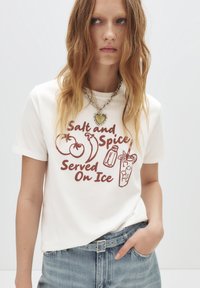 Witte katoenen t-shirt met een grafische print met rode tekst en illustraties van groenten en een drankje. Gecombineerd met lichtblauwe denim.