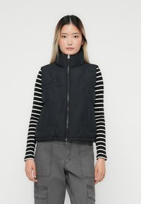 Kvinna i en svart vadderad zip-up väst över en svart-vit randig långärmad tröja och grå cargobyxor, vänd framåt.