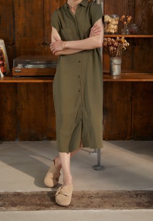 Person i olivengrøn midi-kjole med knapper og beige slip-on sko, stående med armene krydset foran et træhylde med tørrede blomster og en pladespiller.