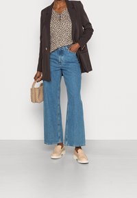 Sort blazer over en mønstret beige bluse, høj talje blå vide jeans og beige loafers. Bærer en lille vævet håndtaske.