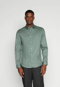 Camicia verde a maniche lunghe realizzata in tessuto morbido; presenta un colletto con bottoni e bottoni frontali. Si adatta bene al corpo ed ha un orlo dritto.