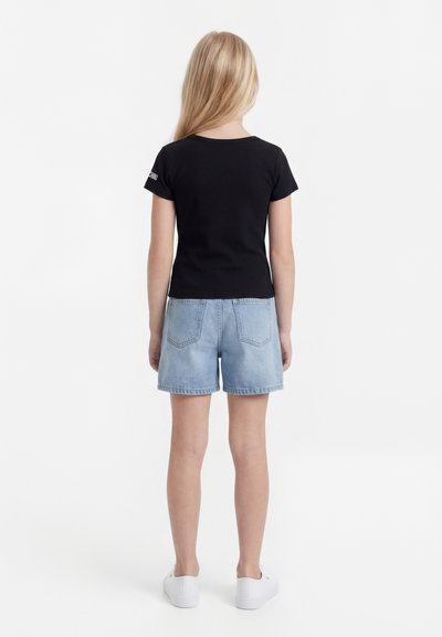 Pikkade blondide juustega tüdruk seisab seljaga vaataja poole, tal on seljas must T-särk, hele sinised teksapüksid ja valged tennised valgel taustal.