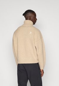 Beige geribbelde trui met hoge kraag, met een klein wit logo op de achterkant en een ontspannen pasvorm.