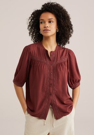 Kvinde med krøllet hår iført en rustfarvet skjorte med knapper og blondedetaljer samt cremefarvede bukser, stående med hænderne i lommerne.