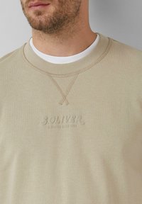 Béžová mikina s rebrovaným okrúhlym výstrihom a tkanou textúrou. Obsahuje vyvýšené logo "SOLIVER" a "ELEVATED SINCE 1969" v zodpovedajúcej farbe.