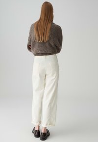 Maglione marrone con texture indossato con pantaloni beige a vita alta con risvolti e due tasche posteriori. Scarpe nere con suola piatta.