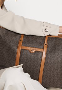 Bras en manteau beige tenant un grand sac fourre-tout Michael Kors marron avec des anses en cuir beige et un motif imprimé du logo.