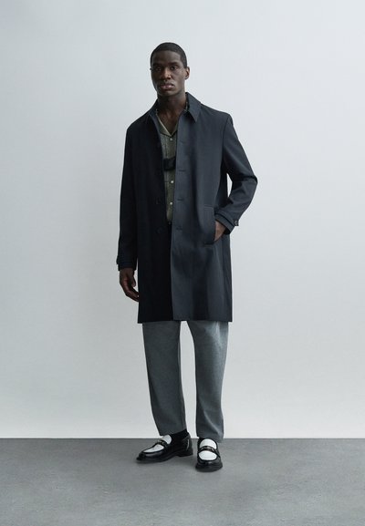 Άνδρας με σκούρο παλτό, γκρι παντελόνια, πουκάμισο με σχέδια και ασπρόμαυρα μοκασίνια στέκεται μπροστά σε απλό γκρι φόντο.