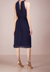 Marinblå, veckad klänning med hög krage och dragkedja bak, instoppad midja, i kombination med metallic guld högklackade skor.