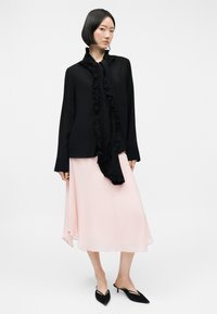 Mujer con cabello corto y negro que lleva una blusa negra de manga larga con volantes y una falda midi rosa claro con zapatos negros de tacón pequeño y puntiagudo.