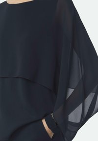 Marineblaue Bluse mit fließenden, transparenten Ärmeln, geschichteter Gestaltung und elastischen Bündchen. Sanfte Textur, leichtes Material, elegante Silhouette.