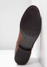 Semelle de chaussure en cuir marron avec grip en caoutchouc noir texturé indiquant la taille 37, positionnée verticalement contre des surfaces blanches.