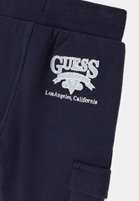 Pantalon de survêtement bleu marine avec ceinture côtelée et logo Guess brodé blanc et bleu clair, ainsi que le texte "Los Angeles California" sur la cuisse.