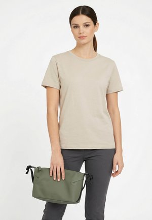 Femme avec une queue de cheval foncée portant un t-shirt beige à manches courtes et un pantalon gris foncé, tenant un sac bandoulière vert avec des bretelles noires.