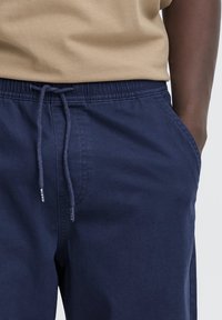 Pantaloni jogger bleumarin cu talie elastică și șnur, prevăzuți cu buzunare laterale și textură netedă. Material din amestec de bumbac.
